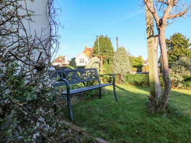 A metal bench on grass next to a tree and bushes with houses in the background at Alcombe House Minehead