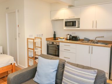 A compact kitchen with white cabinets microwave oven toaster kettle and a sink next to a gray sofa with blue and patterned cushions at The Mew in Dartmouth