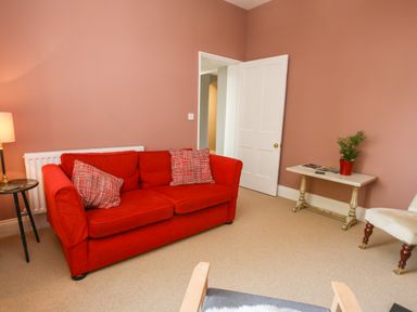 A living room with a red sofa and cushions a lamp on a side table a white door an antique table with a plant and a chair at Gable Lodge in Malvern
