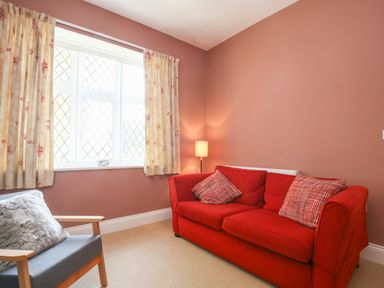 A room with a red sofa with two patterned cushions a grey armchair with a grey cushion a floor lamp and a window with curtains at Gable Lodge in Malvern