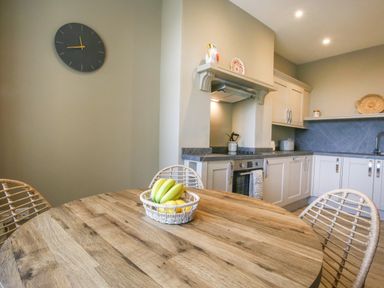A kitchen with a round wooden table and wicker chairs and a fruit basket on the table at Gable Lodge in Malvern