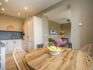 A kitchen with wooden table and chairs and fruit basket opening to a living room with pink sofa and lamp at Gable Lodge in Malvern
