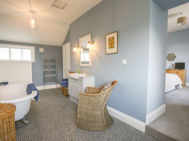 A bathroom with a freestanding bathtub a wicker chair a vanity with a sink and mirror and a doorway leading to a bedroom at Gable Lodge Malvern