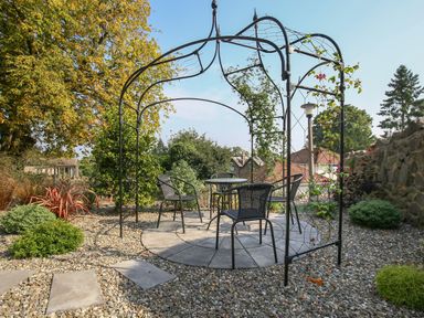 An outdoor seating area with a metal gazebo frame a round table and four chairs surrounded by plants and gravel at Gable Lodge in Malvern