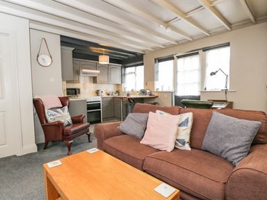 A living area with a brown sofa with cushions a wooden table and a leather armchair next to a kitchen at Paddock Cottage Nr Malmesbury