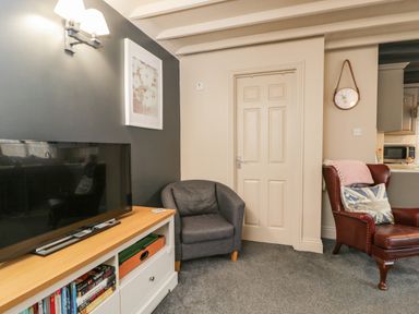 A living room with a television on a white cabinet a gray armchair a brown leather chair with a pillow and blanket a wall clock and a closed door at Paddock Cottage Nr Malmesbury