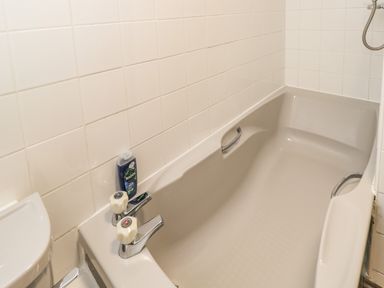 A beige bathtub with two white faucet handles and a bottle of shower gel in a tiled bathroom at Paddock Cottage Nr Malmesbury
