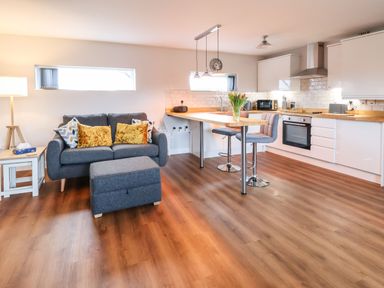 An open plan living room and kitchen with a grey sofa and ottoman a wooden floor and a kitchen island with bar stools at Meols Holiday Lodge in Meols