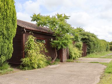 A paved driveway with plants and trees next to a brown wooden building and a gate at Meols Holiday Lodge in Meols