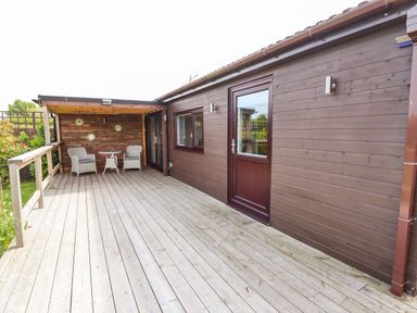 A wooden deck with two chairs and a small table under a roof next to a wooden building at Meols Holiday Lodge in Meols