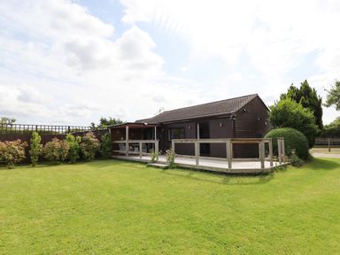 A single-story wooden lodge with a tiled roof and a surrounding wooden deck in a grassy yard with bushes and trees at Meols Holiday Lodge in Meols