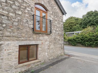 A stone exterior wall with two wooden framed windows next to a roadside with trees and bushes in the background at Coach House in Arnside