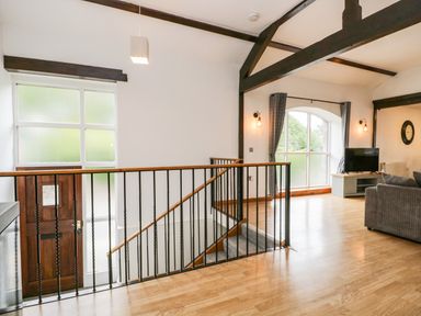 An open living area with a wooden staircase a brown front door large windows and a grey sofa at Coach House in Arnside