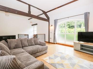 A living room with a large gray sofa a flat screen tv on a cabinet a patterned rug and large window at Coach House in Arnside