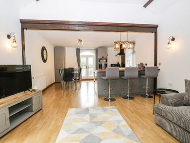 An open plan living area with a gray sofa a television unit a dining table with chairs and a kitchen island with bar stools at Coach House in Arnside