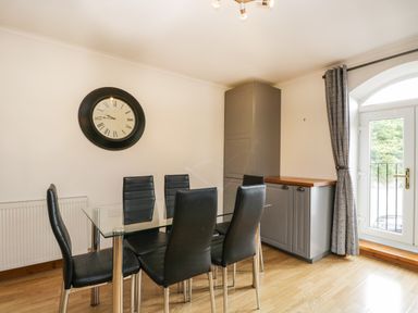 A dining area with a glass table and six black chairs a large clock on the wall gray cabinets and a glass door with gray curtains at Coach House in Arnside