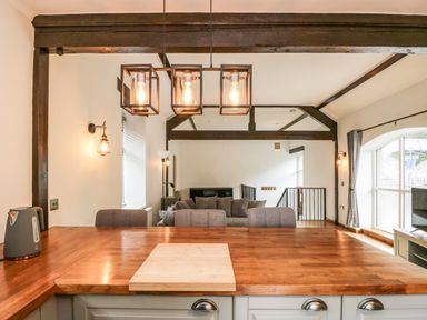 A kitchen countertop with a kettle and a cutting board overlooking a living area with gray sofas and wooden beams at Coach House in Arnside