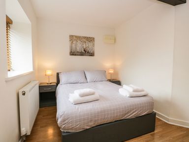 A bedroom with a double bed covered with grey checkered bedding two bedside tables with lamps a radiator and a painting on the wall at Coach House in Arnside