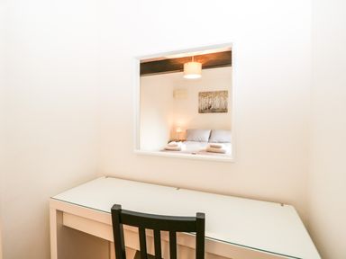A small desk with a black chair in front of a window looking into a bedroom with a bed and lamp at Coach House in Arnside