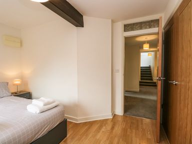 A bedroom with a bed, bedside table with lamp, towels on the bed, wooden floor, and a door opening to a hallway with stairs at Coach House in Arnside