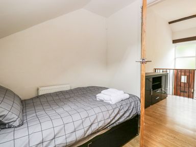 A small bedroom with a single bed and gray grid patterned bedding and a wooden door open to a hallway with wooden flooring and a black cabinet at Coach House Arnside