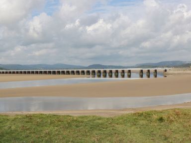 A bridge over sand and water with grass in the foreground and hills in the background at Coach House in Arnside