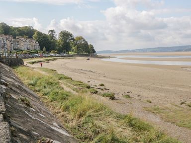 A sandy beach with grassy areas and a row of buildings and parked cars along the road at Coach House in Arnside