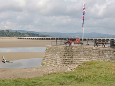 A stone wall with benches and flagpole near a sandy beach with a bird flying at Coach House in Arnside