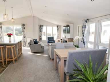 A living room and dining area with grey chairs a wooden table a kitchen island with stools and floral curtains at Dershel Lodge in Southerness
