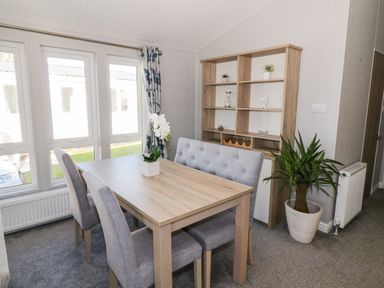 A dining area with a wooden table grey upholstered chairs a wooden shelving unit and a potted plant at Dershel Lodge in Southerness