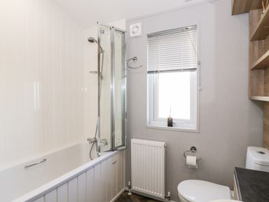 A bathroom with a bathtub shower combination a window a toilet a radiator and shelves at Dershel Lodge in Southerness