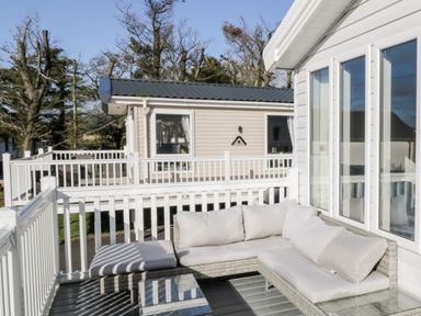 An outdoor patio area with white cushioned wicker sofa and glass table surrounded by white railings at Dershel Lodge in Southerness