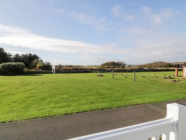 A green lawn next to a paved pathway with trees and bushes in the background at Dershel Lodge in Southerness