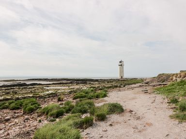 A rocky coastal area with patches of grass and a lighthouse in the distance at Dershel Lodge in Southerness