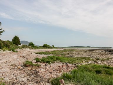 A rocky shoreline with patches of grass and bushes near hills at Dershel Lodge in Southerness