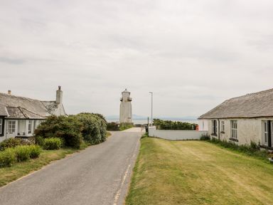 A road leading to a lighthouse between two white stone buildings with grass and bushes at Dershel Lodge in Southerness