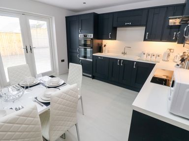 A kitchen with black cabinets and white countertops next to a dining table with white chairs at LunaRoxx 6 Meadow Gardens Kilgetty