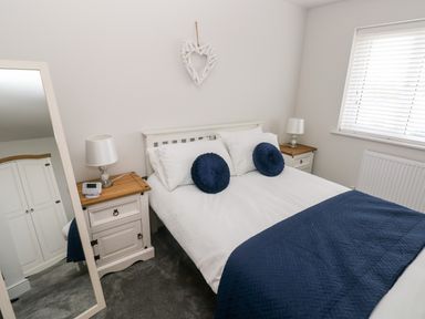 A bedroom with a bed with white bedding and blue cushions beside a window and two bedside tables with lamps at LunaRoxx 6 Meadow Gardens Kilgetty