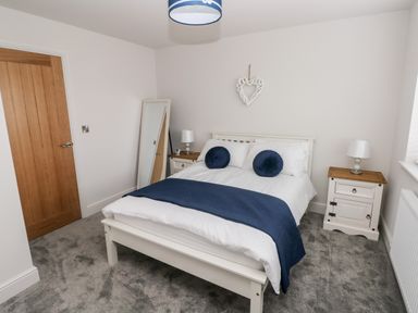 A bedroom with a white bed with blue pillows and a blue throw a wooden door two white bedside tables with lamps a full-length mirror on a grey carpet a heart-shaped wall decoration at LunaRoxx 6 Meadow Gardens Kilgetty