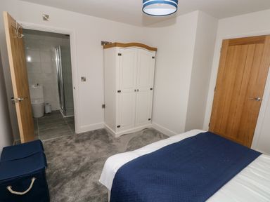 A bedroom with a bed covered by a blue blanket a white wardrobe a blue laundry basket and a door leading to a bathroom at LunaRoxx 6 Meadow Gardens Kilgetty