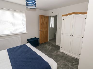 A bedroom with a bed covered in white and blue bedding a white wardrobe with wooden top a wooden door partially open showing a bathroom and a window with white blinds at LunaRoxx 6 Meadow Gardens Kilgetty