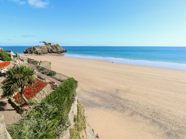 A beach with a sandy shore and a small island with a building in the sea seen from a walkway with plants and benches at LunaRoxx 6 Meadow Gardens Kilgetty