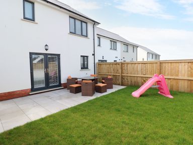 A backyard with a grass lawn a pink slide and a patio area with patio furniture at LunaRoxx 6 Meadow Gardens Kilgetty
