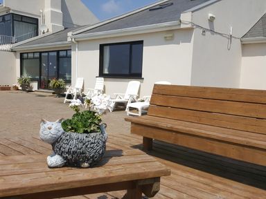 An outdoor seating area with a wooden bench and table with a cat-shaped flower pot and white flowers in front of a white building with white chairs at High Tide in Duncannon County Wexford