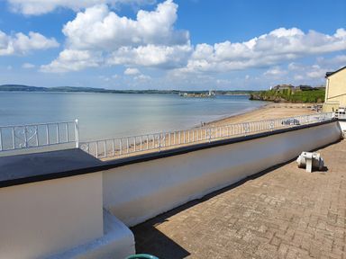 A seaside view with a sandy beach and a ship on the water behind a white wall with metal railings at High Tide in Duncannon County Wexford