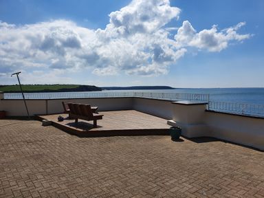 An outdoor patio area with a wooden bench on a wooden deck overlooking the sea with a stone wall and railing at High Tide in Duncannon County Wexford