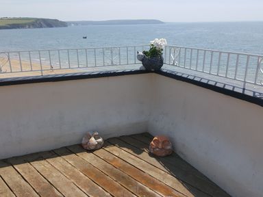 A wooden deck with two stone face sculptures on the floor and a cat-shaped flower pot with white flowers on a white railing with an ocean view at High Tide in Duncannon County Wexford