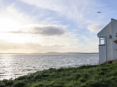 A grassy area next to a body of water with a white house on the right side at Beach House in Duncannon County Wexford