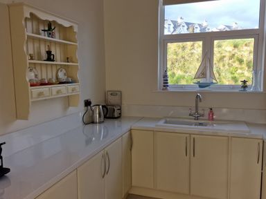 A kitchen with white cupboards a countertop a sink a window with decorative items and a shelf with cups and plates at Beach House in Duncannon County Wexford
