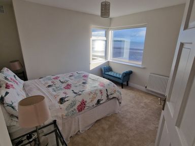 A bedroom with a double bed with floral bedding a glass bedside table with lamps a blue upholstered bench under corner windows showing a sea view at Beach House in Duncannon County Wexford